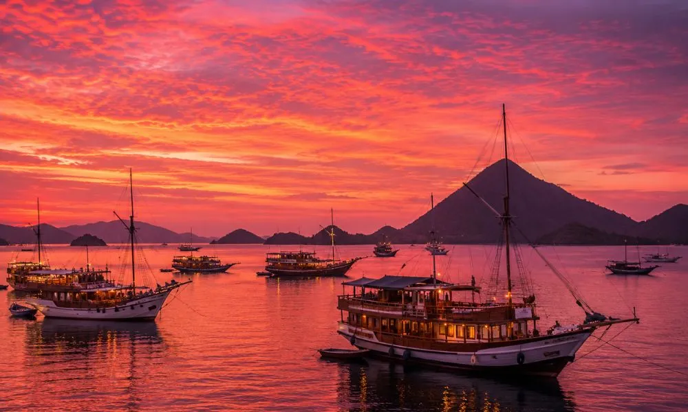 A deeply saturated pink and orange sunset over Labuan Bajo harbour in Flores, Indonesia, with traditional wooden pinisi boats silhouetted against volcanic mountains in the background