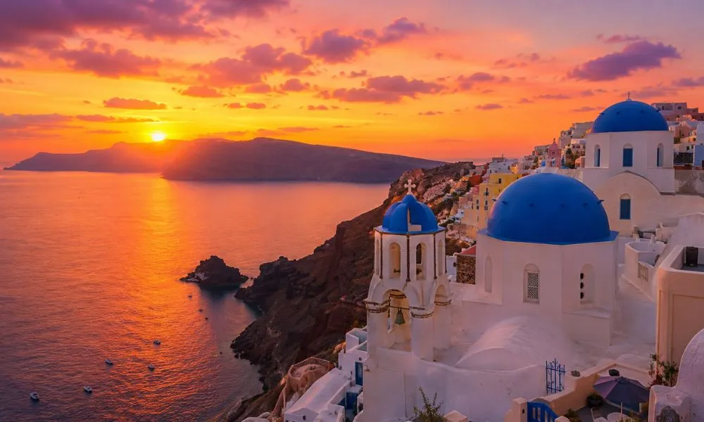 Golden sunset over Oia village in Santorini, Greece, featuring iconic white-washed buildings with blue-domed churches cascading down the volcanic cliffside above the Aegean Sea